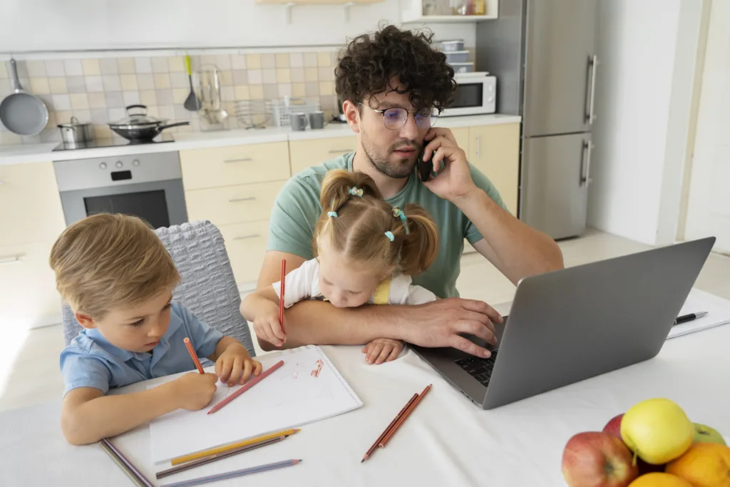 A multitasking dad on a call and using his laptop at the table while two kids happily draw nearby