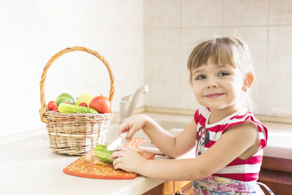 Smiling little girl in the kitchen, cutting vegetables with a butter knife