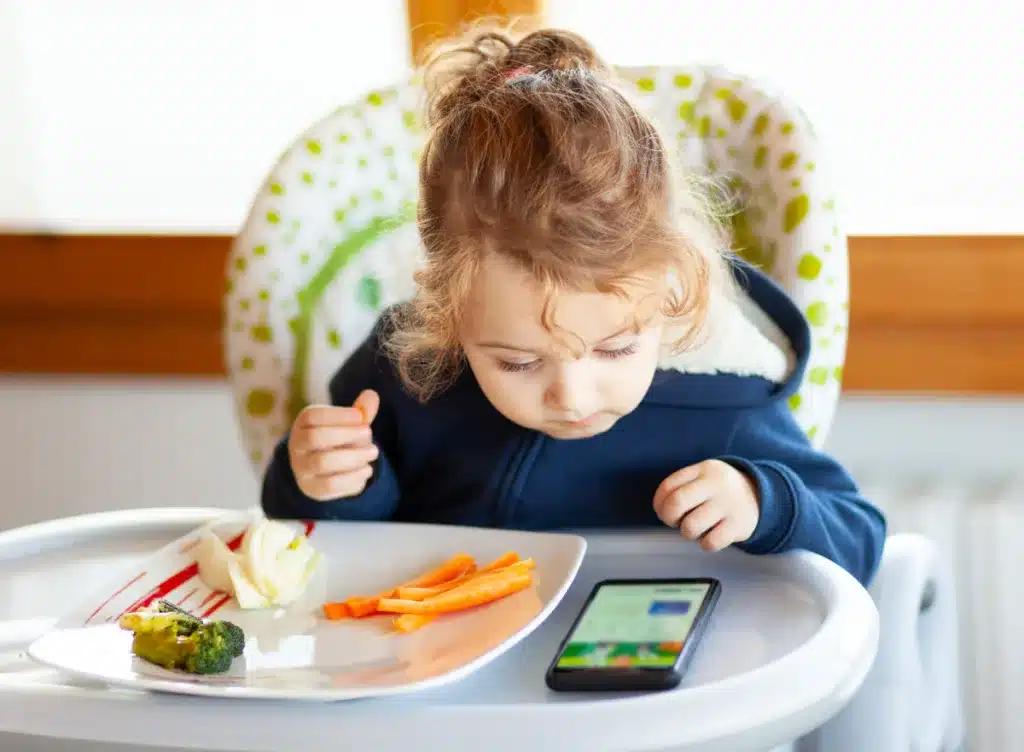Toddler in high chair eating while watching cartoons on phone