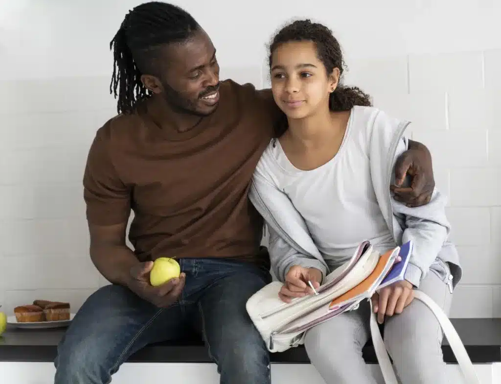 A proud looking father smiles while sitting on a bench with his arm wrapped around his smiling daughter by his side