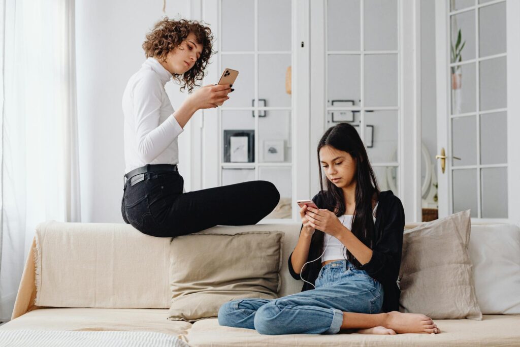Two teenage girls sitting on a sofa, absorbed in their phones, scrolling through content.