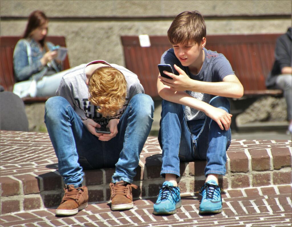 Two school-aged boys sitting side by side, each engrossed in their own smartphone.
