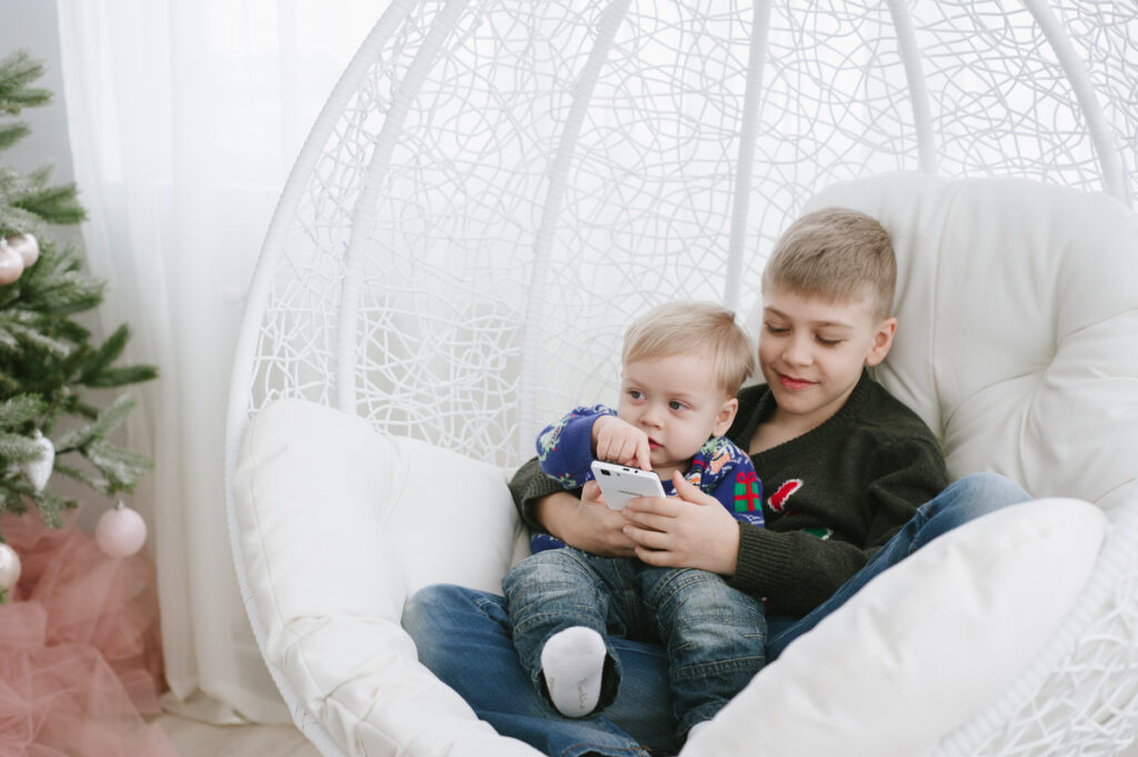 Little brother glides on big brother’s phone while they both sit comfortably in a nest chair
