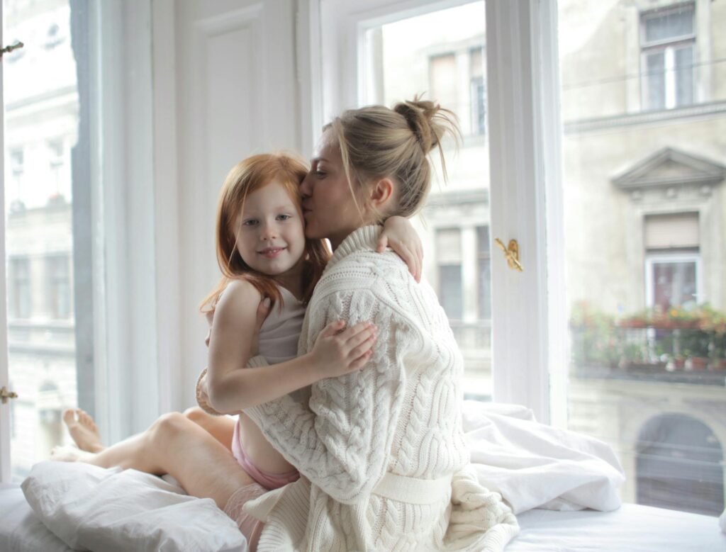 Mother sitting on bed, holding daughter in her arms, sharing a tender moment together.