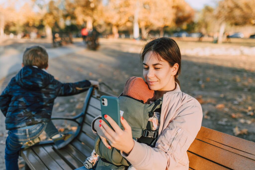 Mother sitting on bench with two children beside her, looking at phone screen while spending time outdoors.