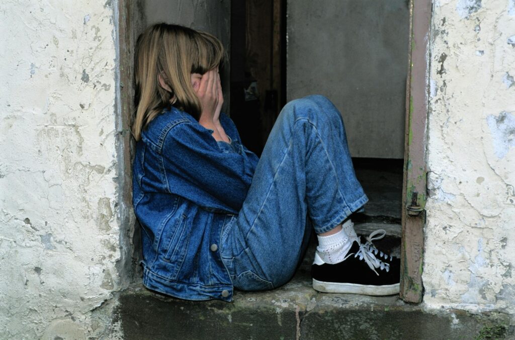 A eight-year-old girl sits in a corner, crying with her face covered by her hands.