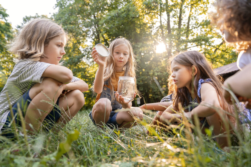 School-aged kids in the forest sitting on the grass observing an insect in a jar