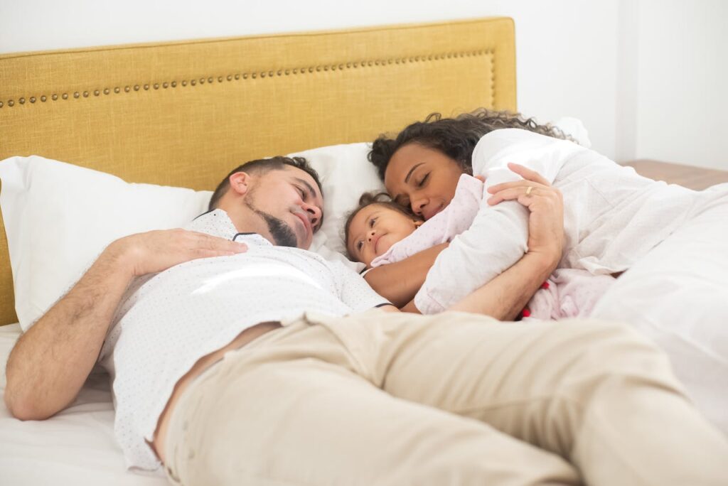 A mother and father pretend to sleep in bed, hoping to encourage their little child beside them to fall asleep by mimicking their behavior.