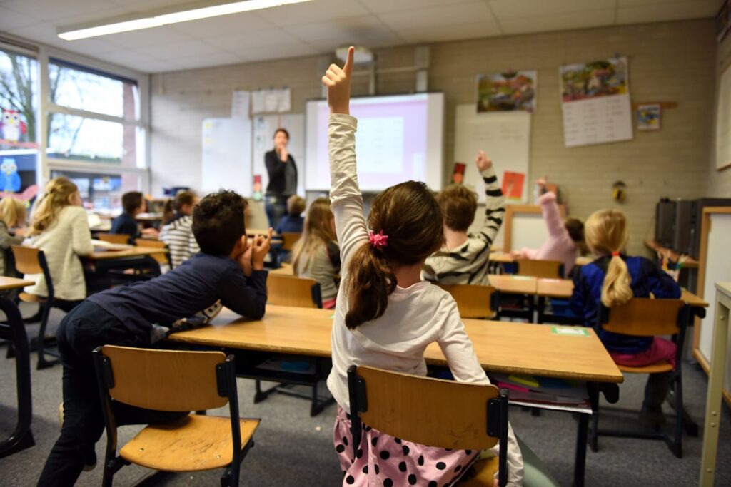 View from the back of a classroom showing kids standing and raising their hands.